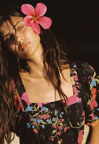 A woman with long dark hair and a pink flower on her head, wearing a black floral top with pink, blue, and yellow flowers against a dark background.