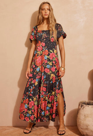 A woman modeling a black maxi dress with a vibrant red, pink, and blue floral print, standing against a textured beige wall.