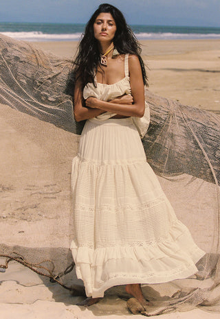 A woman stands on a sandy beach wearing a long cream maxi dress with a tiered ruffle skirt. A large beige fishing net is draped across the beach behind her, with the ocean and blue sky in the background.