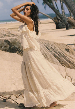 A woman wearing a flowing cream boho-style maxi dress stands on a sandy beach next to a large driftwood log, with palm trees and the ocean in the background.